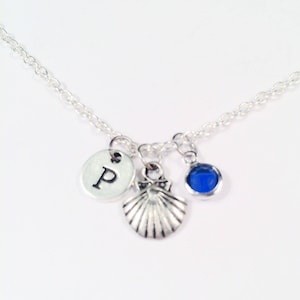 Op de afbeelding: Een zilveren ketting met een schelpbedel, een blauwe edelsteenbedel en een zilveren bedel met de letter "P" erop.