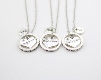 May include: Three silver necklaces with heart pendants that say "Big Sister", "Middle Sister", and "Little Sister". Each pendant has a small rhinestone and is surrounded by a circle with an inscription. The necklaces have initial charms.