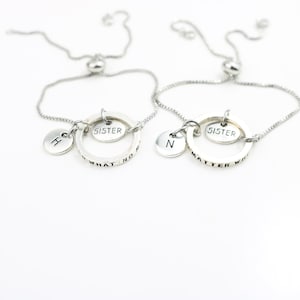 Op de afbeelding: Twee zilveren vriendschapsarmbanden met verstelbare koorden. Elke armband heeft een bedel met het woord "Zuster" en een bedel met een letter, een met de letter "H" en de andere met de letter "N". De armbanden hebben ook een ringbedel met de woorden "Wat er ook gebeurt" en "Wat ook maar".