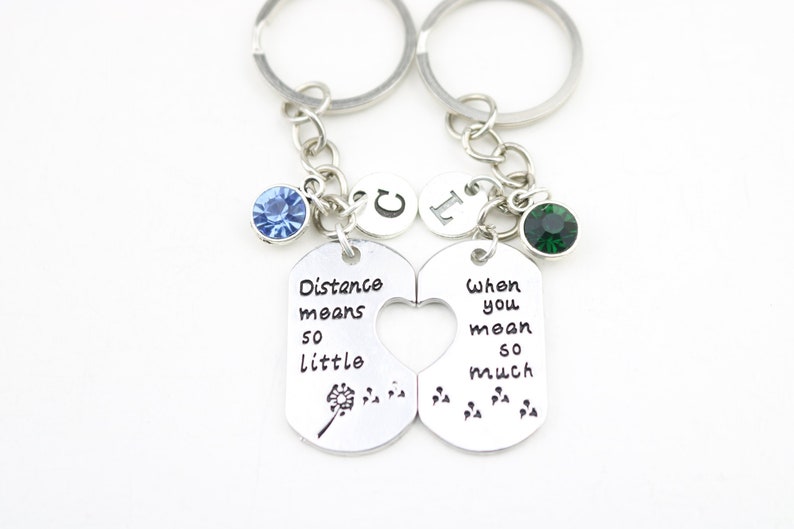 May include: Two silver keychains with a heart-shaped cutout. One keychain has the text "Distance means so little" with a dandelion design. The other keychain has the text "When you mean so much" with a footprint design. Both keychains have a blue and green crystal charm.