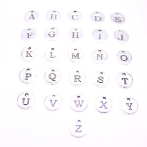 May include: A set of 26 silver metal alphabet letter charms. Each charm is a circle with a single letter printed in black.