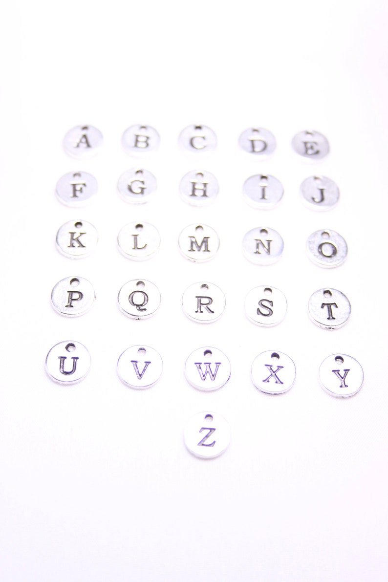 May include: A set of 26 silver metal alphabet letter charms. Each charm is a circle with a single letter printed in black.