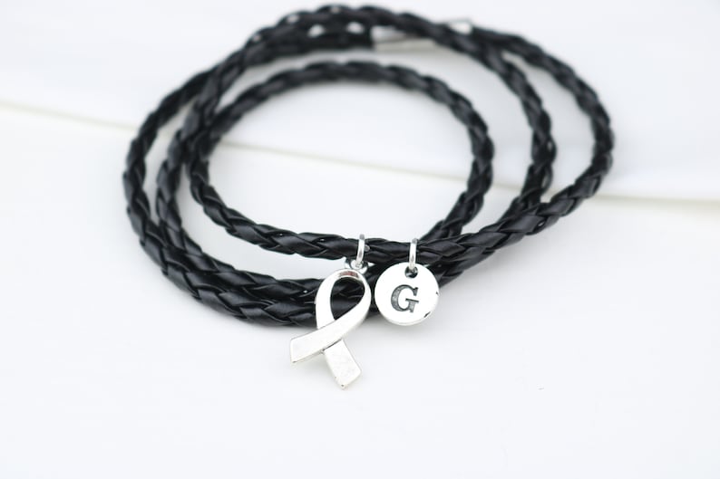 May include: A black leather braided bracelet with a silver ribbon charm and a silver charm with the letter "G" engraved on it.