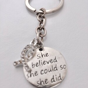 May include: Silver keychain with a round charm that says "She believed she could so she did". The charm has a small rhinestone embellishment.