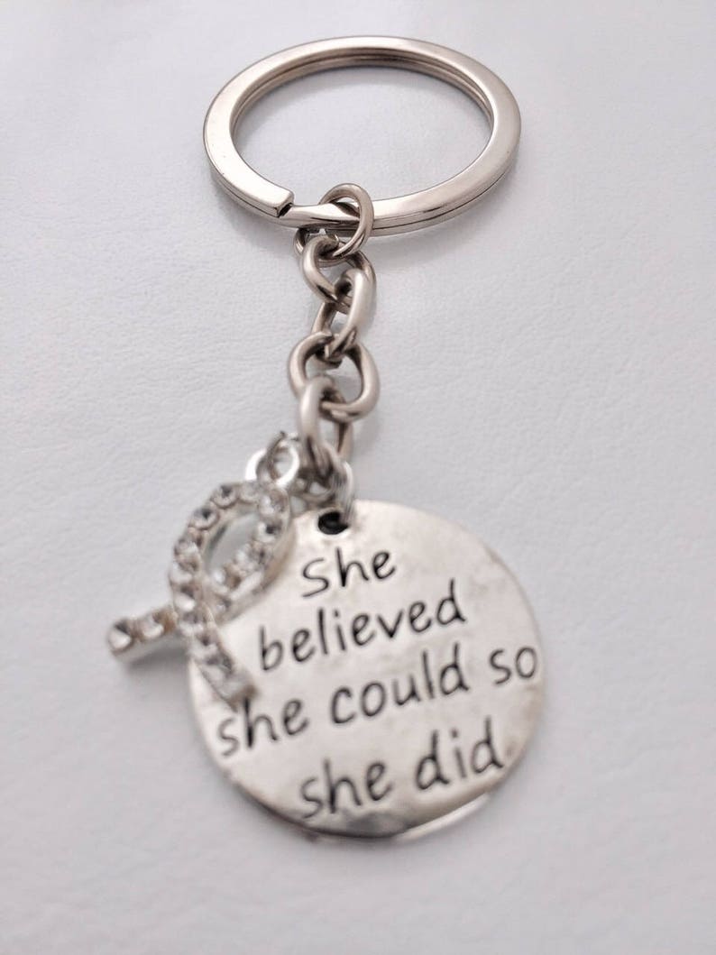 May include: Silver keychain with a round charm that says "She believed she could so she did". The charm has a small rhinestone embellishment.