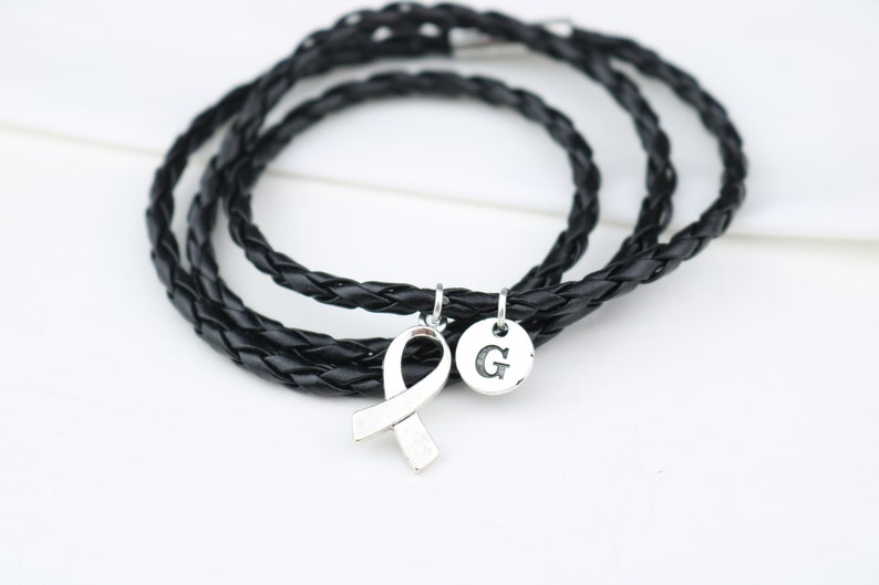 May include: A black leather braided bracelet with a silver ribbon charm and a silver charm with the letter "G" engraved on it.