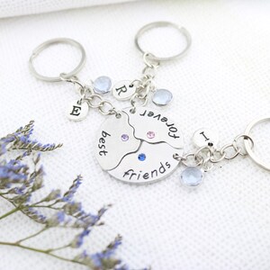 May include: Silver keychains with a 'best friends forever' design. The keychains are connected to a circular pendant split into three parts, each with a gem. Individual charms with letters and gems are also attached.