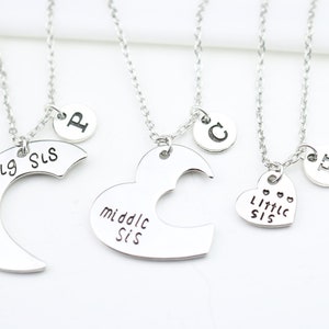 May include: Three silver necklaces with heart and crescent moon pendants. The pendants have the words "Big Sis", "Middle Sis", and "Little Sis" engraved on them. Each necklace also has a small charm with a letter on it.