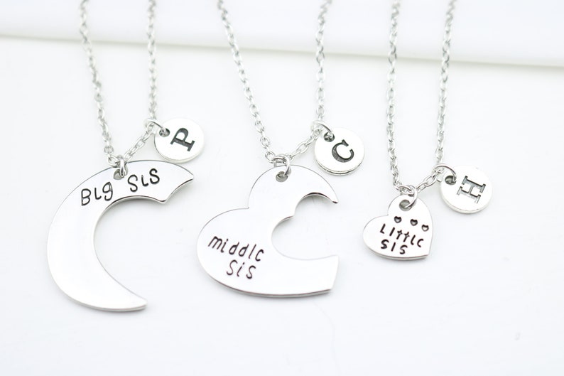 May include: Three silver necklaces with heart and crescent moon pendants. The pendants have the words "Big Sis", "Middle Sis", and "Little Sis" engraved on them. Each necklace also has a small charm with a letter on it.