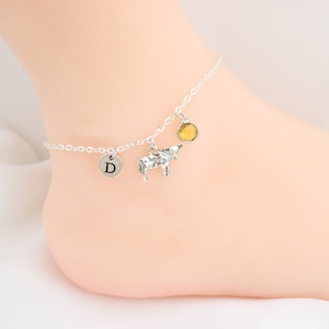May include: Silver ankle bracelet with a cow charm, a letter 'D' charm, and a yellow gemstone charm.