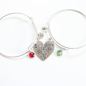 Könnte beinhalten: Zwei silberne Armbänder mit einem geteilten Herz-Anhänger, auf dem "Big Sis" und "Little Sis" steht. Die Anhänger sind mit bunten Schmetterlingen verziert. Die Armbänder haben auch rote und grüne Edelstein-Anhänger.