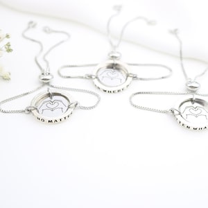 May include: Three silver friendship bracelets with adjustable sliding knots. Each bracelet features a round charm with a hand gesture forming a heart and an inscription. The inscriptions read "No Matter", "Where", and "Then What".