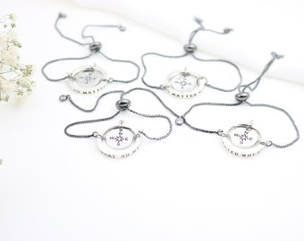 Puede incluir: Cuatro pulseras de plata con cordones ajustables negros. Cada pulsera presenta una brújula con un círculo plateado alrededor y las palabras "No matter what" o "Where will" inscritas en el círculo.