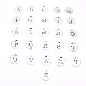 May include: A set of 26 silver metal alphabet letter charms. Each charm is a circle with a single letter printed in black.