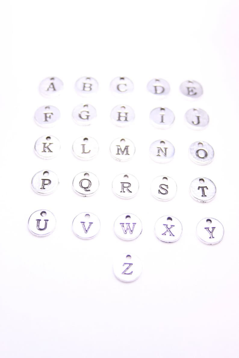 May include: A set of 26 silver metal alphabet letter charms. Each charm is a circle with a single letter printed in black.