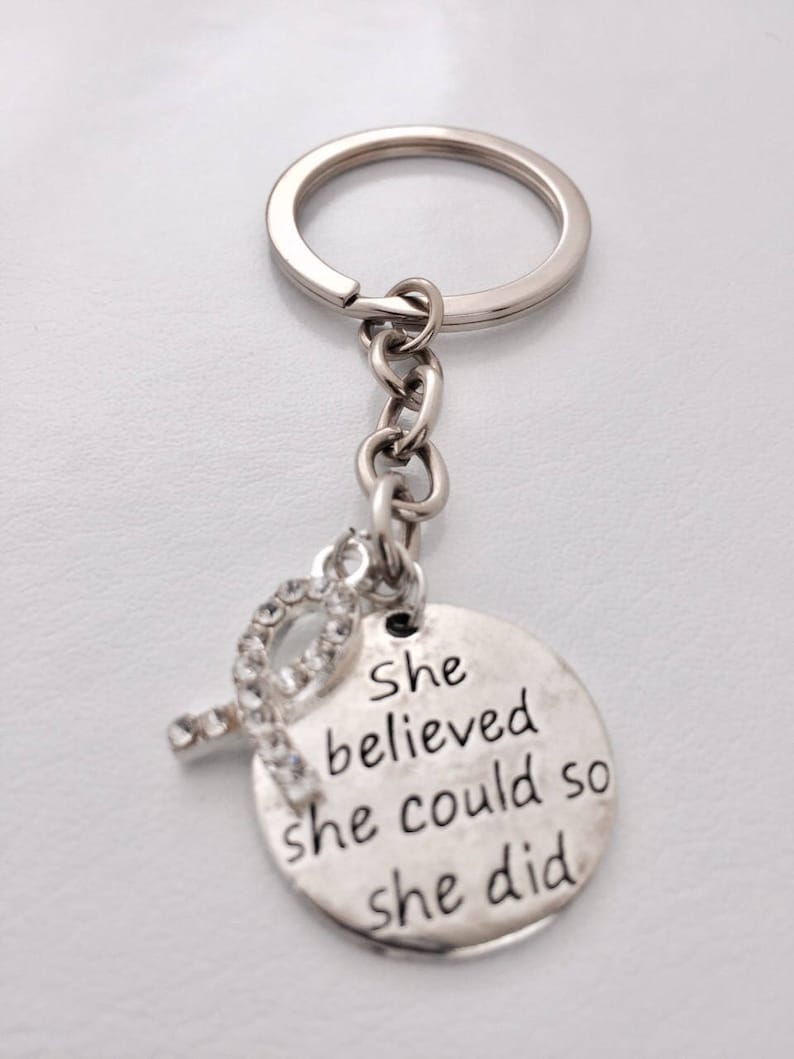 May include: Silver keychain with a round charm that says "She believed she could so she did". The charm has a small rhinestone ribbon attached to it.