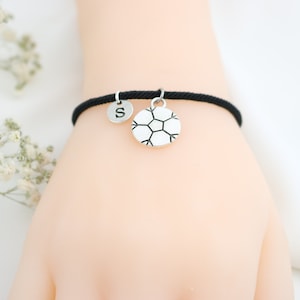 Puede incluir: Pulsera de cordón negro con un dije de balón de fútbol plateado y un dije plateado con la letra "S".