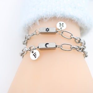以下が含まれることがあります： 太陽のシンボルと「H」の文字が刻まれたチャームが付いたシルバーチェーンブレスレット2本。もう一方のチャームには月のシンボルと「F」の文字が刻まれています。