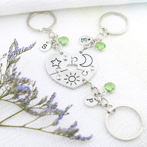 May include: Silver heart-shaped puzzle piece keychains with initials and green accents. One keychain features a star, sun, and moon design. The keychains are connected with a chain and a key ring. A sprig of purple flowers is in the background.
