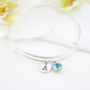 May include: A silver expandable bangle bracelet with a charm that has the letter 'A' and a light blue crystal.