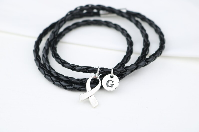 May include: A black leather braided bracelet with a silver ribbon charm and a silver charm with the letter "G" engraved on it.