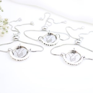 May include: Four silver friendship bracelets with a sliding bead closure. Each bracelet features a circular charm with a line drawing of two hands holding each other. The charms are engraved with the text "No matter where we go", "No matter what we do", "No matter where we are", and "No matter what we face".
