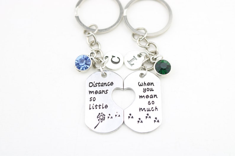 May include: Two silver keychains with a heart-shaped cutout. The left keychain has a blue rhinestone, a dandelion, and the text "Distance means so little". The right keychain has a green rhinestone and the text "When you mean so much".