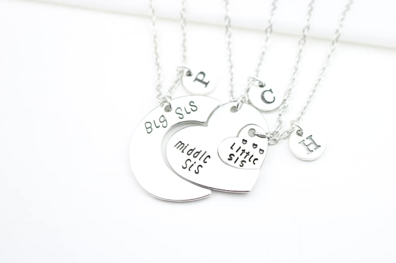 May include: Three silver necklaces with heart and circle pendants. The pendants have the words "Big Sis", "Middle Sis", and "Little Sis" engraved on them. Each necklace also has a small circle pendant with a letter engraved on it.