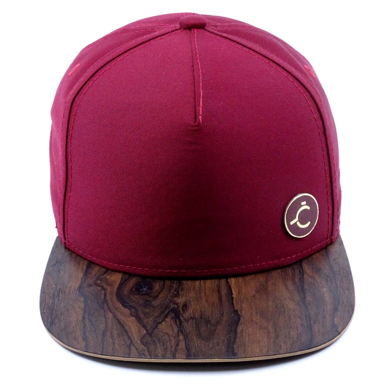 original 5panel burgundy cap with a wooden ziricote palisander visor,on the front is a round logo with our company brand,on the back is a brass buckle with a leather strip on which the company's name is printed,front view