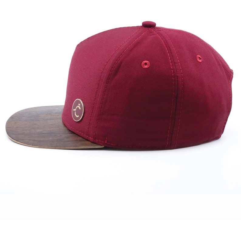 original 5panel burgundy cap with a wooden ziricote palisander visor,on the front is a round logo with our company brand,on the back is a brass buckle with a leather strip on which the company's name is printed,side view