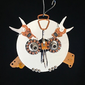 May include: A white owl-shaped ornament made from recycled computer parts. The owl has large, round eyes, a beak, and wings made from circuit boards and other electronic components.