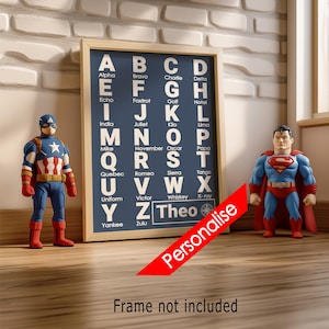 May include: A framed navy blue print with white lettering of the phonetic alphabet, flanked by Captain America and Superman figurines. The print includes the name "Theo". A red banner with the word "Personalise" is across the image. The phrase "Frame not included" is at the bottom.