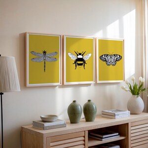 May include: Three framed insect art prints. Each print features a black and white illustration of a dragonfly, bee, and butterfly against a yellow background. The frames are light wood, and the artwork is displayed on a neutral-colored wall.