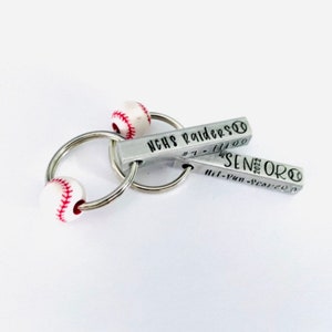 May include: A silver keychain with two baseball-shaped charms. The charms are engraved with the text "NCHS Raiders #1 - 11/09" and "Senior 2022 - 11/09".