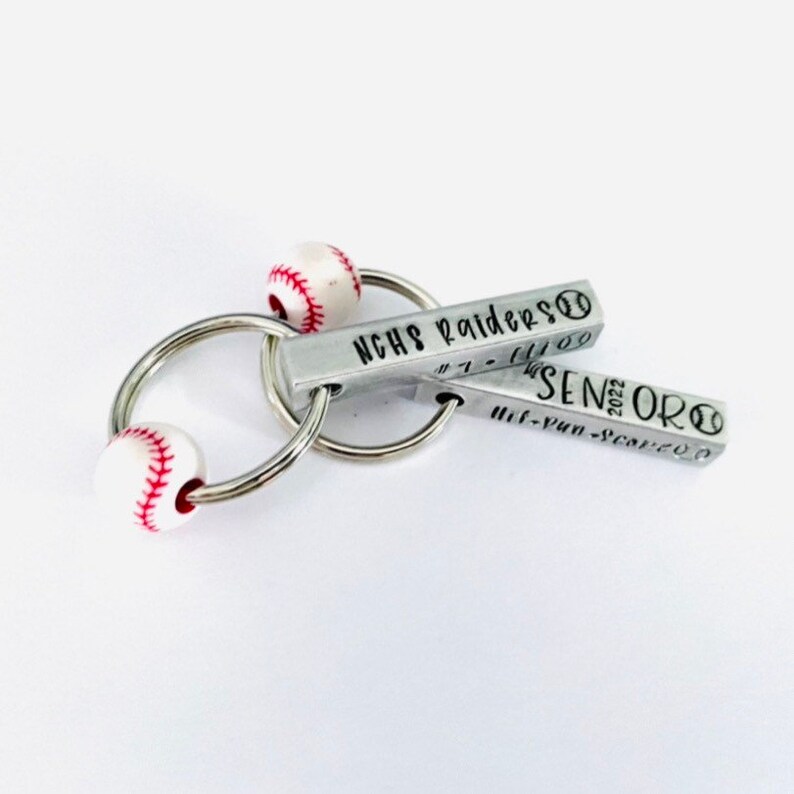 May include: A silver keychain with two baseball-shaped charms. The charms are engraved with the text "NCHS Raiders #1 - 11/09" and "Senior 2022 - 11/09".