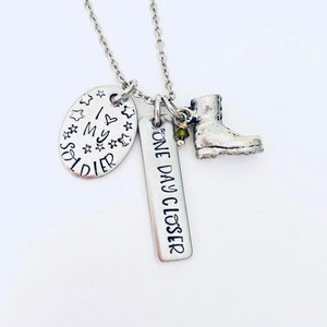 May include: Silver necklace with three charms: a boot, a tag that says "One Day Closer", and a tag that says "I Love My Soldier" with stars around the edge.
