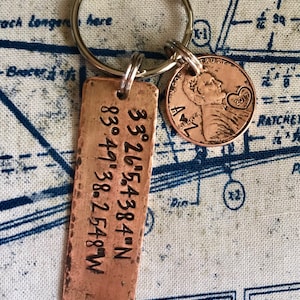 May include: A copper keychain with a rectangular tag engraved with coordinates: 33°26′5.4384"N 83°49′38.2548 W. A copper penny with the year 2017 engraved on it is attached to the keychain.