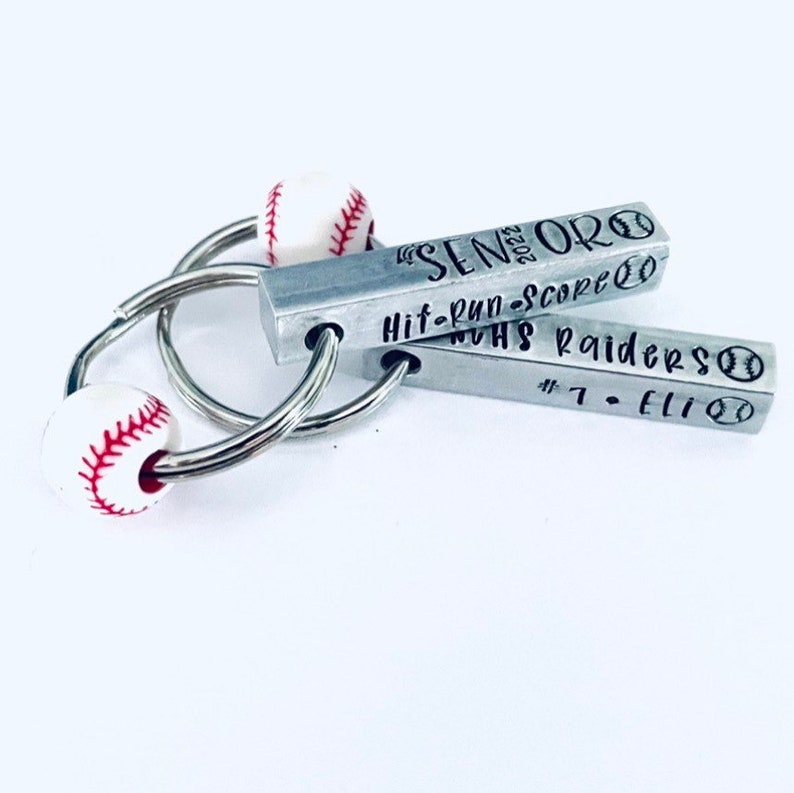 May include: A silver keychain with two metal bars. The top bar has the text "Senior 2023" and "Hit-Run-Score" with a baseball graphic. The bottom bar has the text "NUHS Raiders #7-Eli" with a baseball graphic. The keychain has two white baseball-shaped beads.