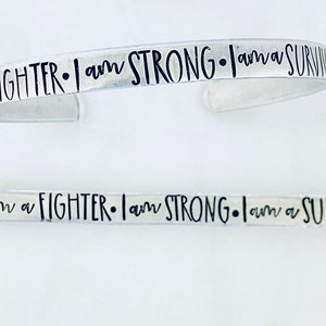 May include: Two silver bracelets with the inscription "I am a FIGHTER. I am STRONG. I am a SURVIVOR." The inscription is in black lettering.