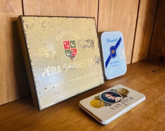 Pode incluir: Conjunto de latas de cigarros vintage em uma superfície de madeira. A maior lata é dourada com escrita árabe e as palavras "Pera Cigarettes London". Uma lata azul Dunhill e uma lata Van Dyck também estão presentes.
