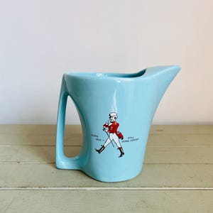 Puede incluir: Jarra de cer&aacute;mica azul claro con un asa y un pico &uacute;nicos. La jarra presenta un gr&aacute;fico de una figura marchando con abrigo rojo, con el texto "BORN 1820 - STILL GOING STRONG".