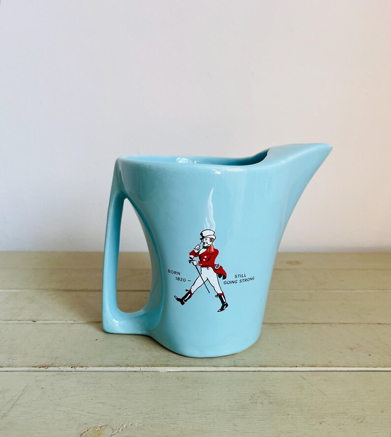 Puede incluir: Jarra de cer&aacute;mica azul claro con un asa y un pico &uacute;nicos. La jarra presenta un gr&aacute;fico de una figura marchando con abrigo rojo, con el texto "BORN 1820 - STILL GOING STRONG".