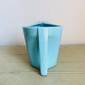 Puede incluir: Una jarra de cer&aacute;mica azul claro con asa. La jarra tiene un dise&ntilde;o angular &uacute;nico y un pico. La jarra est&aacute; sobre una superficie de madera verde claro. El fondo es una pared blanca lisa.