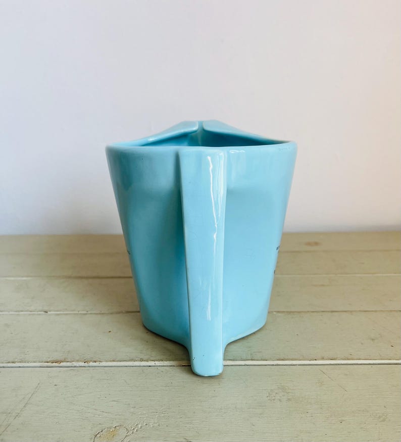 Puede incluir: Una jarra de cer&aacute;mica azul claro con asa. La jarra tiene un dise&ntilde;o angular &uacute;nico y un pico. La jarra est&aacute; sobre una superficie de madera verde claro. El fondo es una pared blanca lisa.