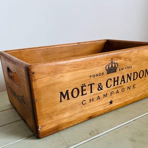 Puede incluir: Caja de champán de madera con la inscripción "MOËT & CHANDON CHAMPAGNE" en negro. La caja es rectangular, con un asa en el lateral. La madera tiene un tono marrón cálido. El texto incluye una corona y una estrella.