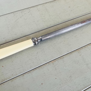 May include: A steel knife sharpener with a cream-coloured handle and a silver bolster. The sharpening steel is long and tapered, with a slightly rusted tip. The handle is smooth and rounded.