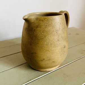 May include: A beige ceramic jug with a rounded body and a handle. The jug has a speckled, textured surface.