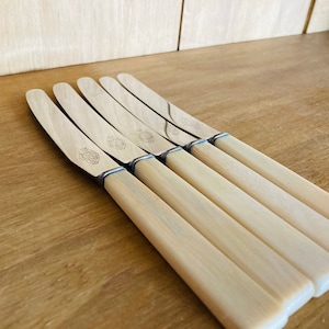 May include: Five silver butter knives with ivory-coloured handles. The knives are arranged in a row, with the handles overlapping.