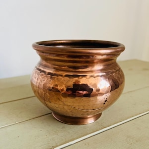 Puede incluir: Un cuenco de cobre martillado con cuerpo redondeado y borde acampanado. El cuenco tiene un color marrón rojizo cálido y una superficie texturizada. Se apoya sobre una pequeña base circular. El interior del cuenco es liso y oscuro.