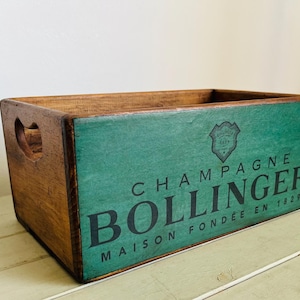 May include: A rectangular wooden crate with a natural brown finish and a teal-painted front panel. The front features black text reading CHAMPAGNE BOLLINGER MAISON FONDÉE EN 1829 and a small crest logo. The crate includes cutout handles on the sides.